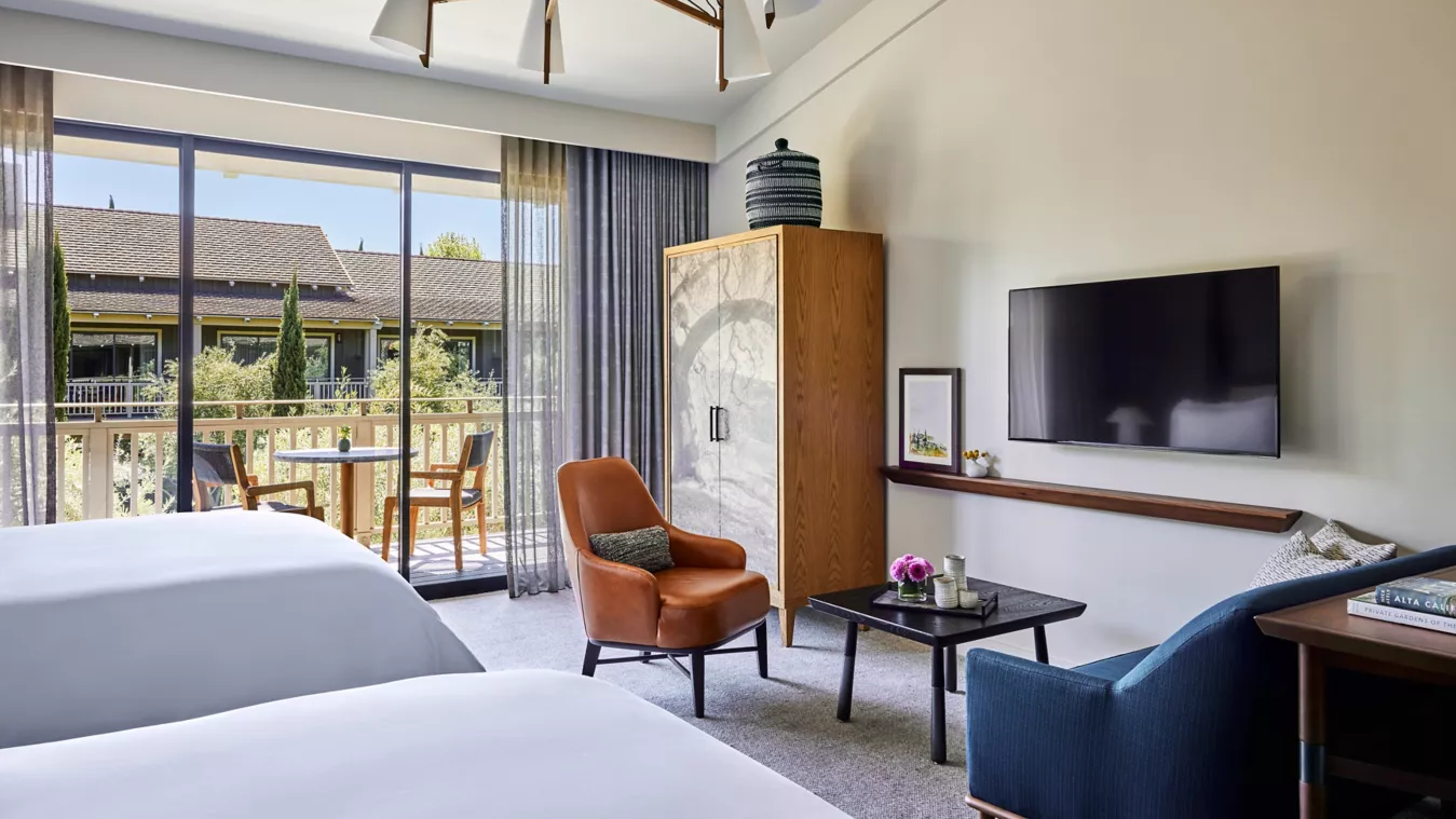 Hotel room with two beds, balcony view, seating area with an orange armchair, and a TV.