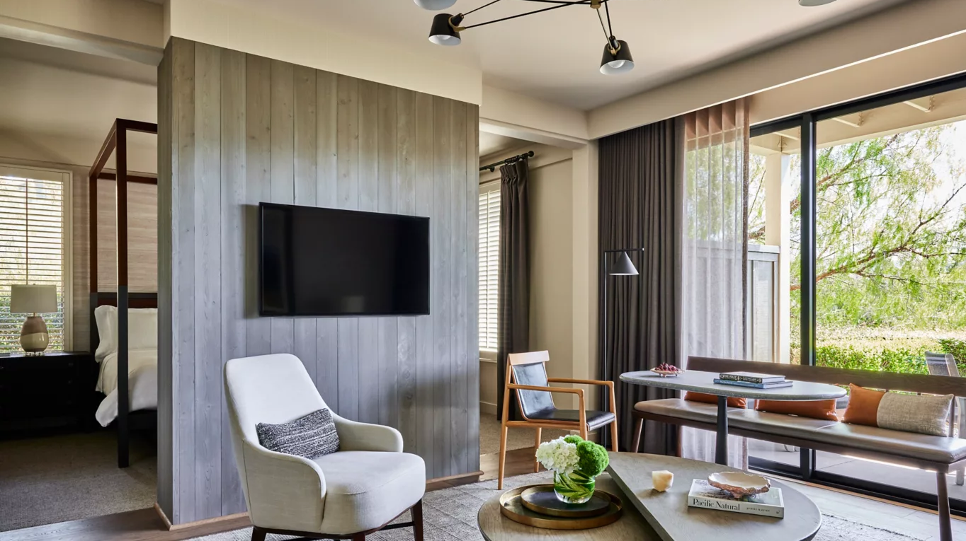 Living area of a hotel suite: chair, TV, table, bench, and bed visible through a divider.