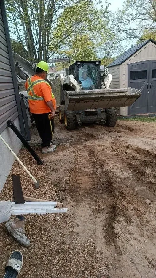 Construction worker in safety vest directs a skid steer loader on a muddy driveway, next to a building and shed.