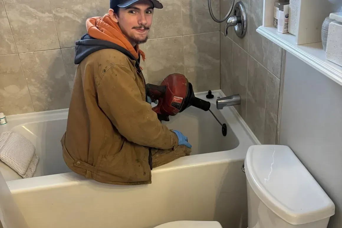 Man in a bathroom uses a drain snake. He sits in a tub with plumbing tools. Smiling.