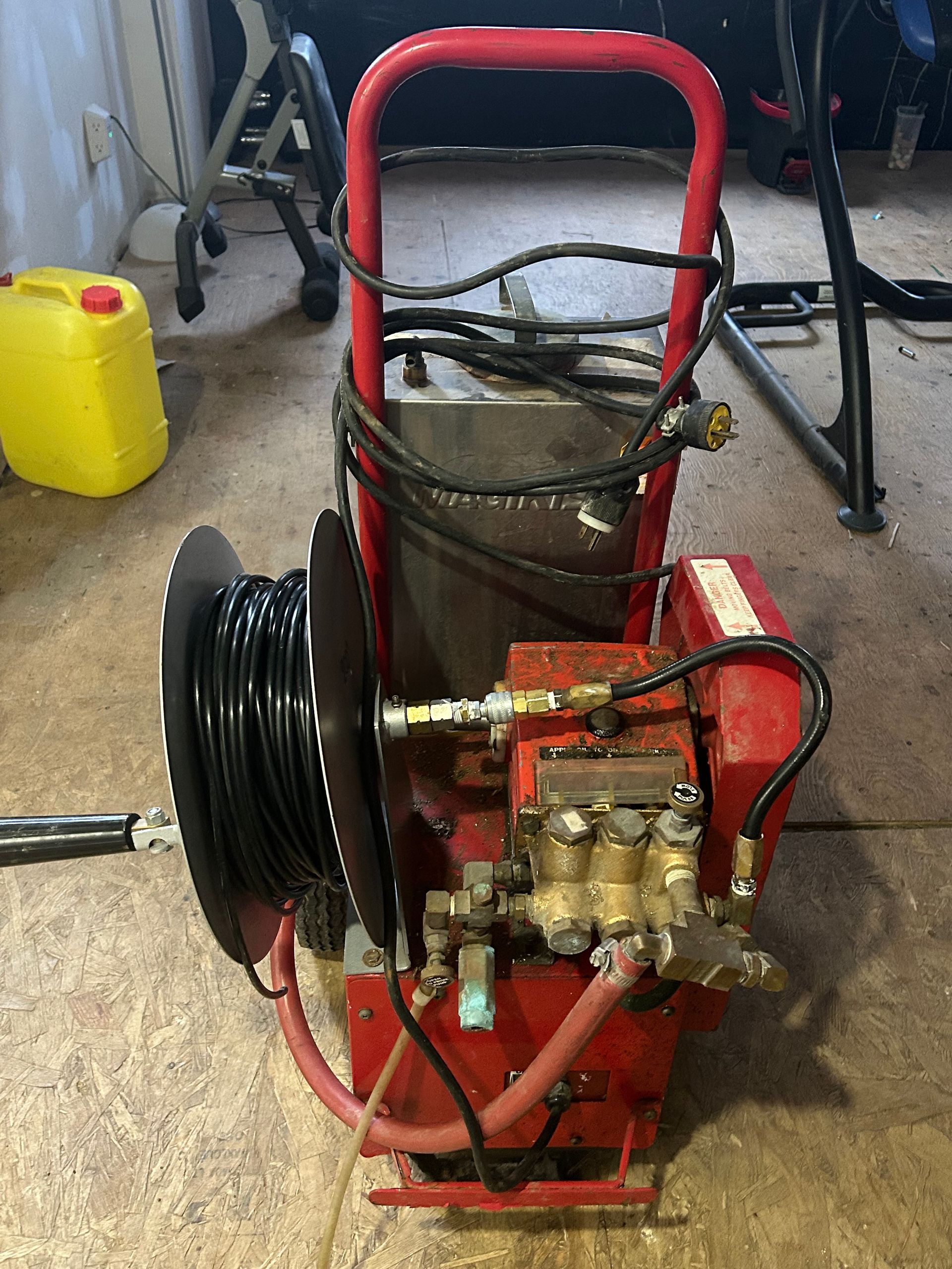 Red and silver sewer jetter machine with a black hose reel. Yellow container in background.