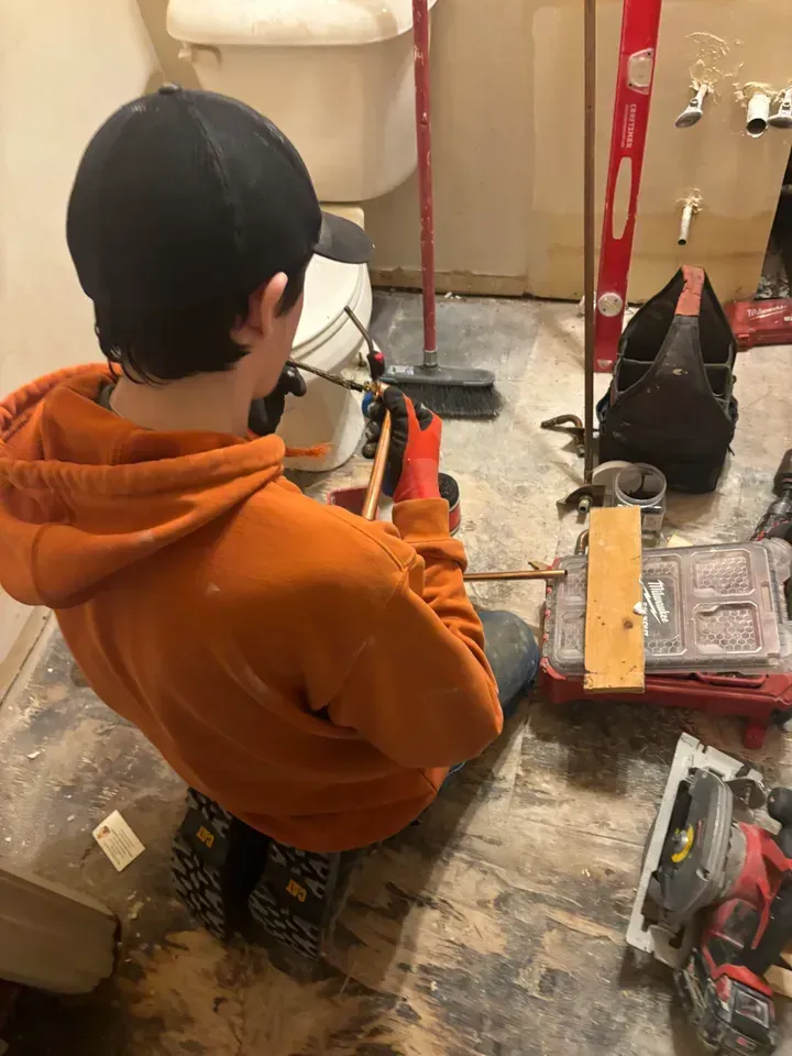 Person in orange hoodie working on plumbing in a bathroom, near a toilet and tools.
