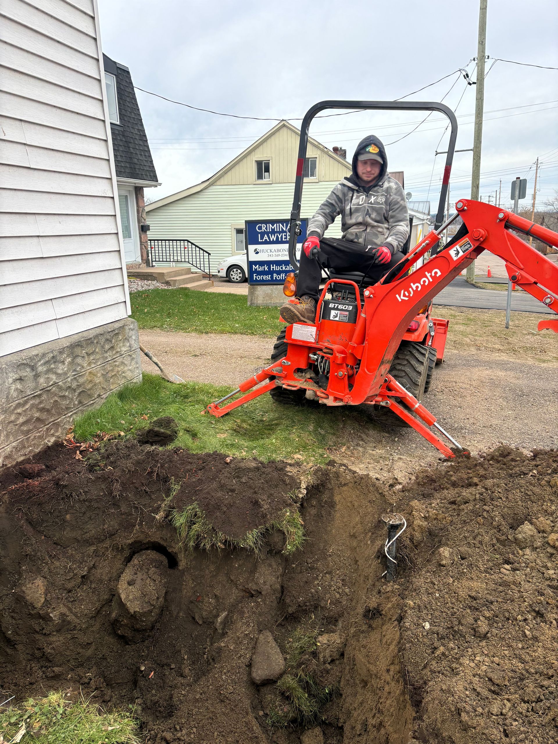 Person operating an orange Kubota backhoe, digging near a building.