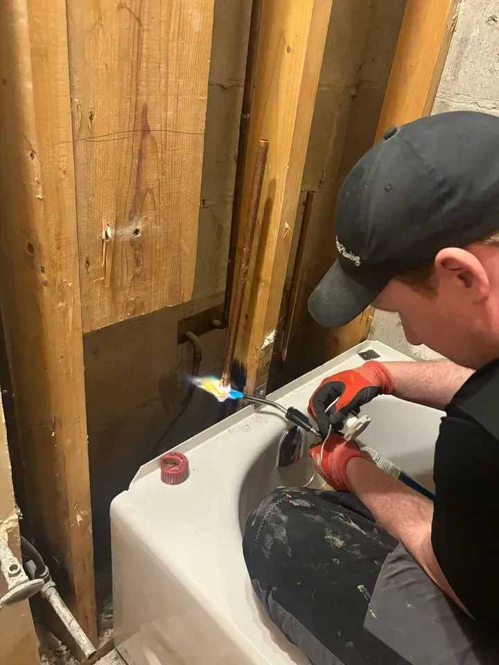 Person using a torch to solder pipes near a bathtub. Wood framing is visible behind him.