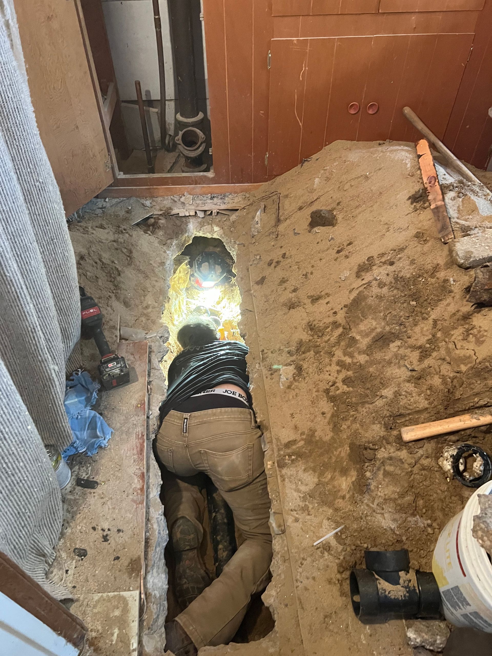 Person in trench-like hole, working on plumbing. Dusty environment, brown pants, black shirt, overhead pipe visible.