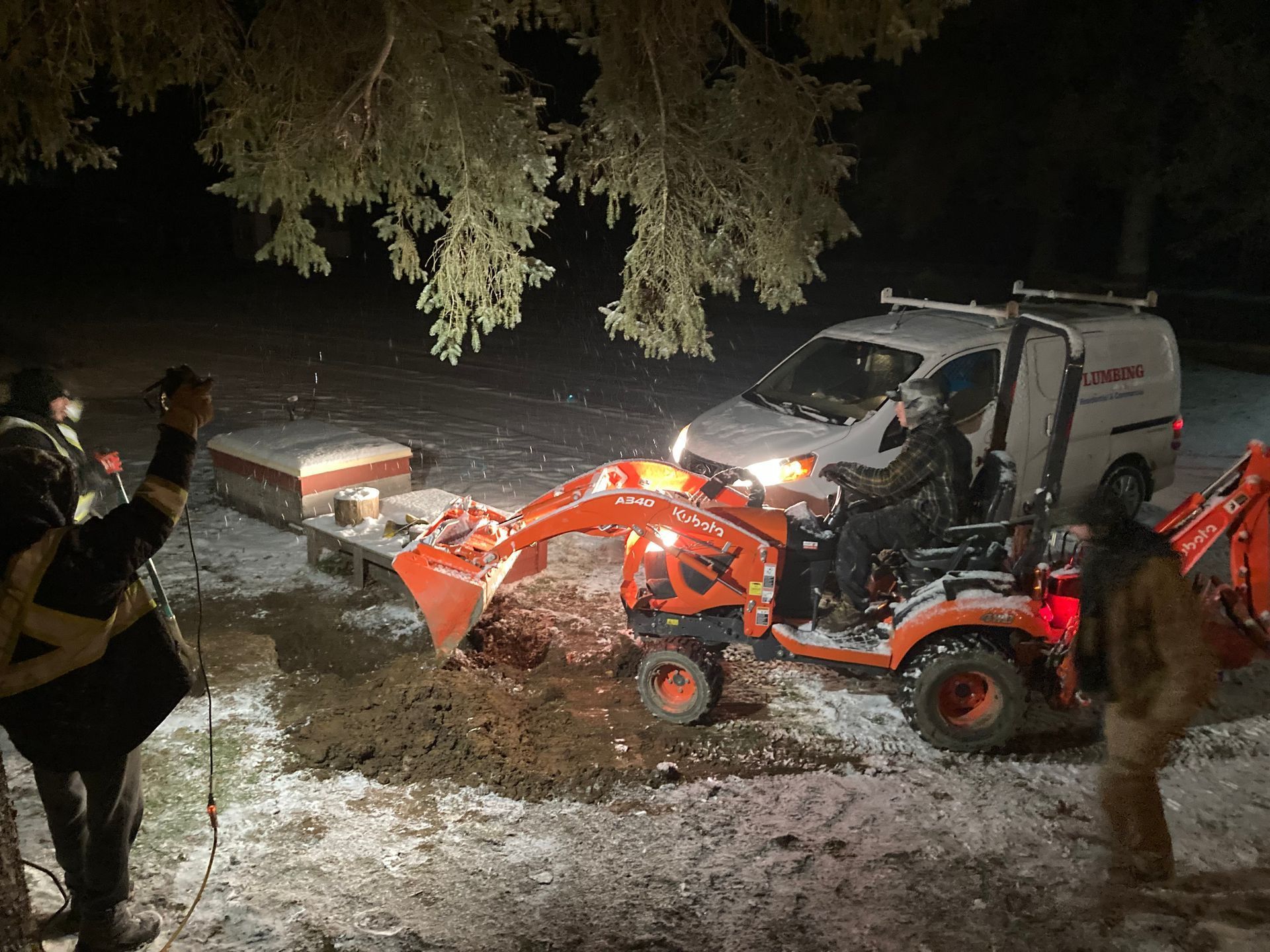 Orange tractor with a front-end loader clearing debris in the snow. A van and people are nearby at night.