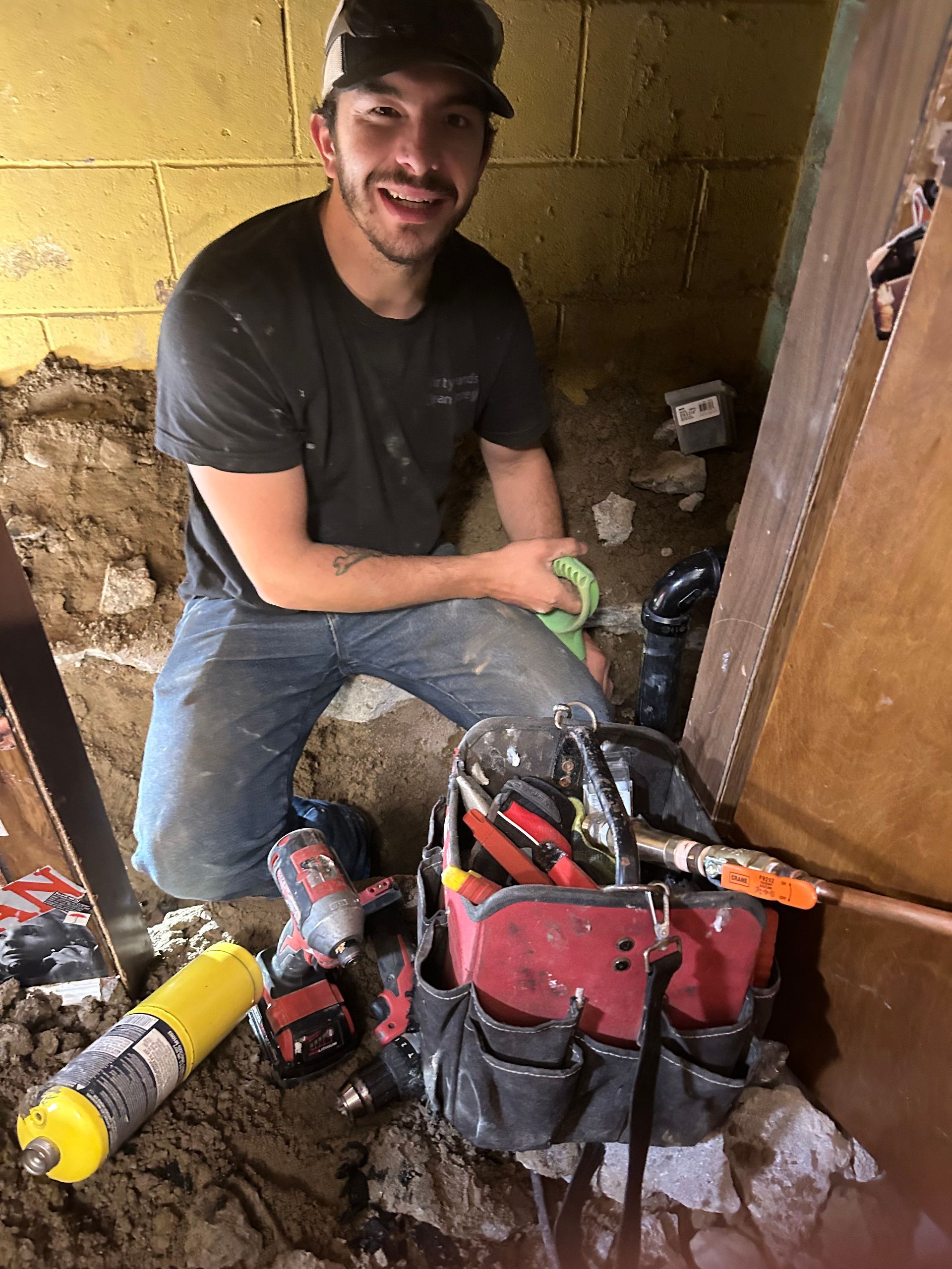 Smiling worker kneels amidst tools, fixing pipes in a dimly lit, damp space.