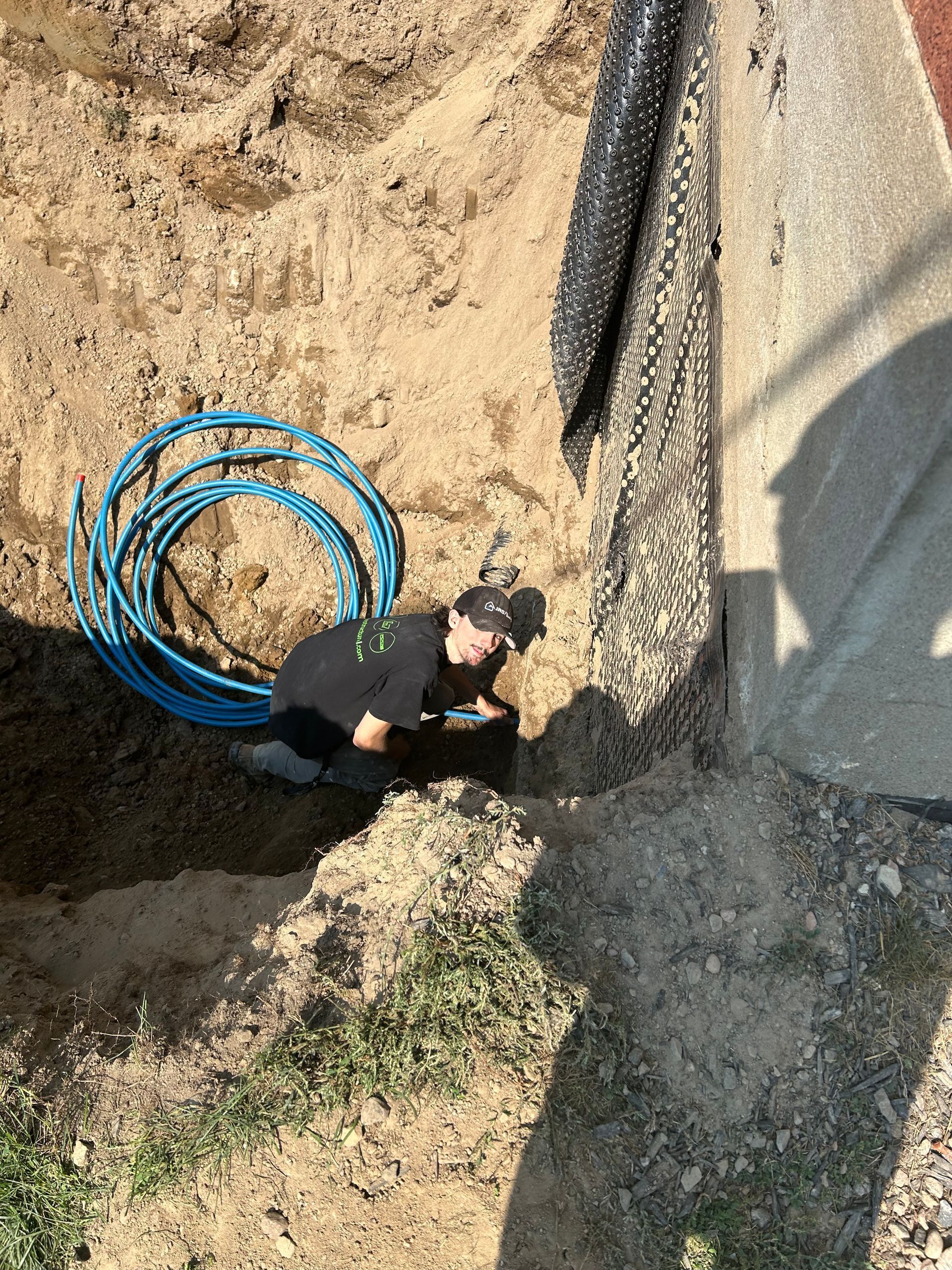 A person in a ditch works near coiled blue tubing and a wall. Sunlight casts shadows.