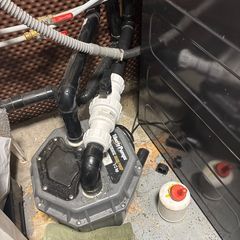 Basement sump pump with black pipes, white valves, gray housing, and a red/yellow lid.