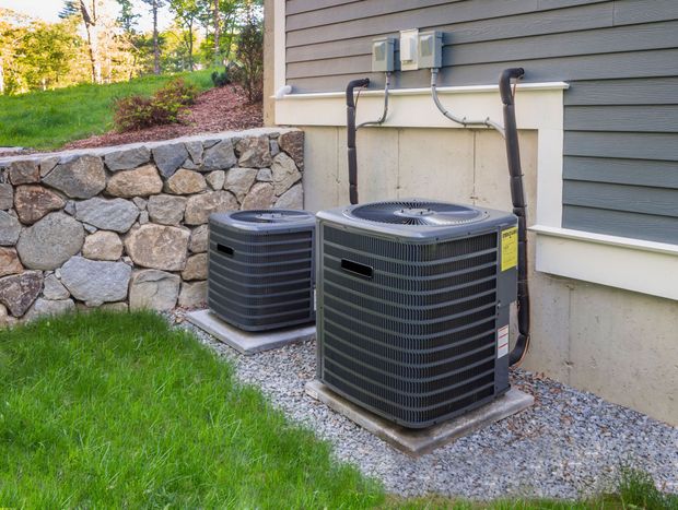 Two black air conditioning units outside a house, next to a stone wall and green grass.