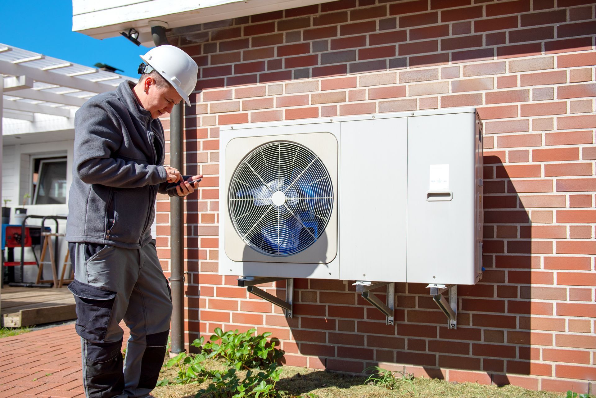 HVAC technician inspecting outdoor AC unit on brick wall during maintenance or installation work.