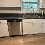 Kitchen with white cabinets, stainless dishwasher, black sink, window, and light wood floor.