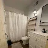 Small bathroom with white shower curtain, toilet, sink vanity, mirror, and wall shelves.