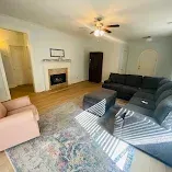 Bright living room with fireplace, dark sofa and chairs, rug, and ceiling fan.