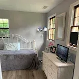 Bright bedroom with white bed, gray bedding, dresser with TV, and windows letting in daylight