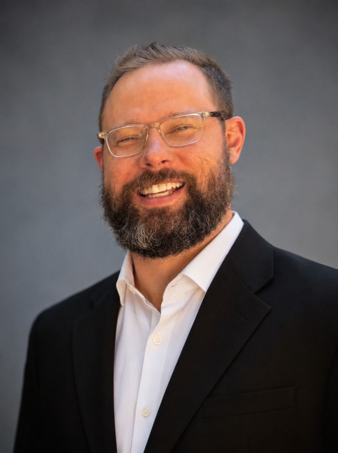 Smiling man with glasses and beard in a black blazer and white shirt against a gray background Smiling man with glasses and beard in a black blazer and white shirt against a gray background