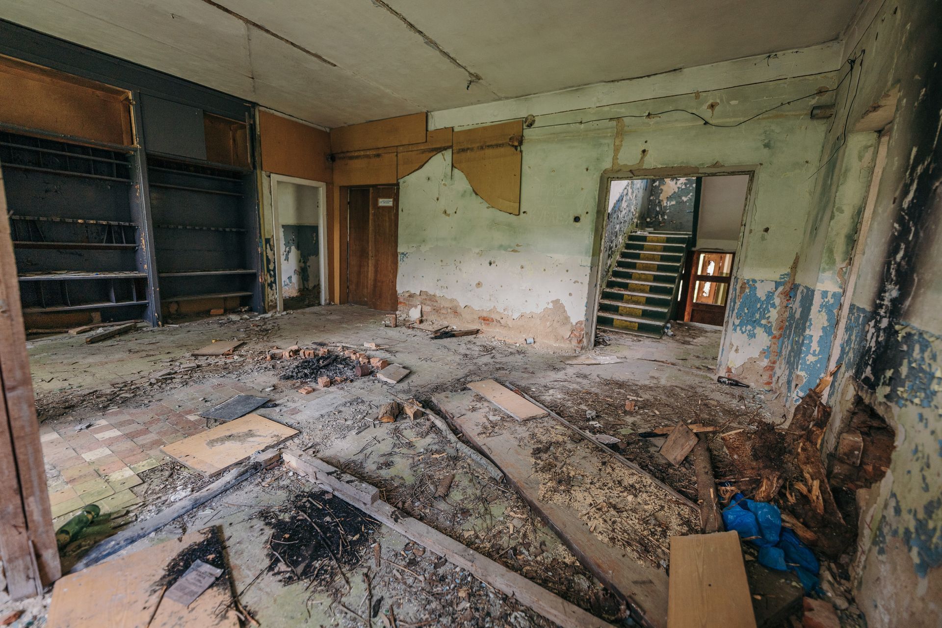Flood-damaged room with debris, peeling walls, broken floor, and exposed shelving near a stairwell. Flood-damaged room with debris, peeling walls, broken floor, and exposed shelving near a stairwell.