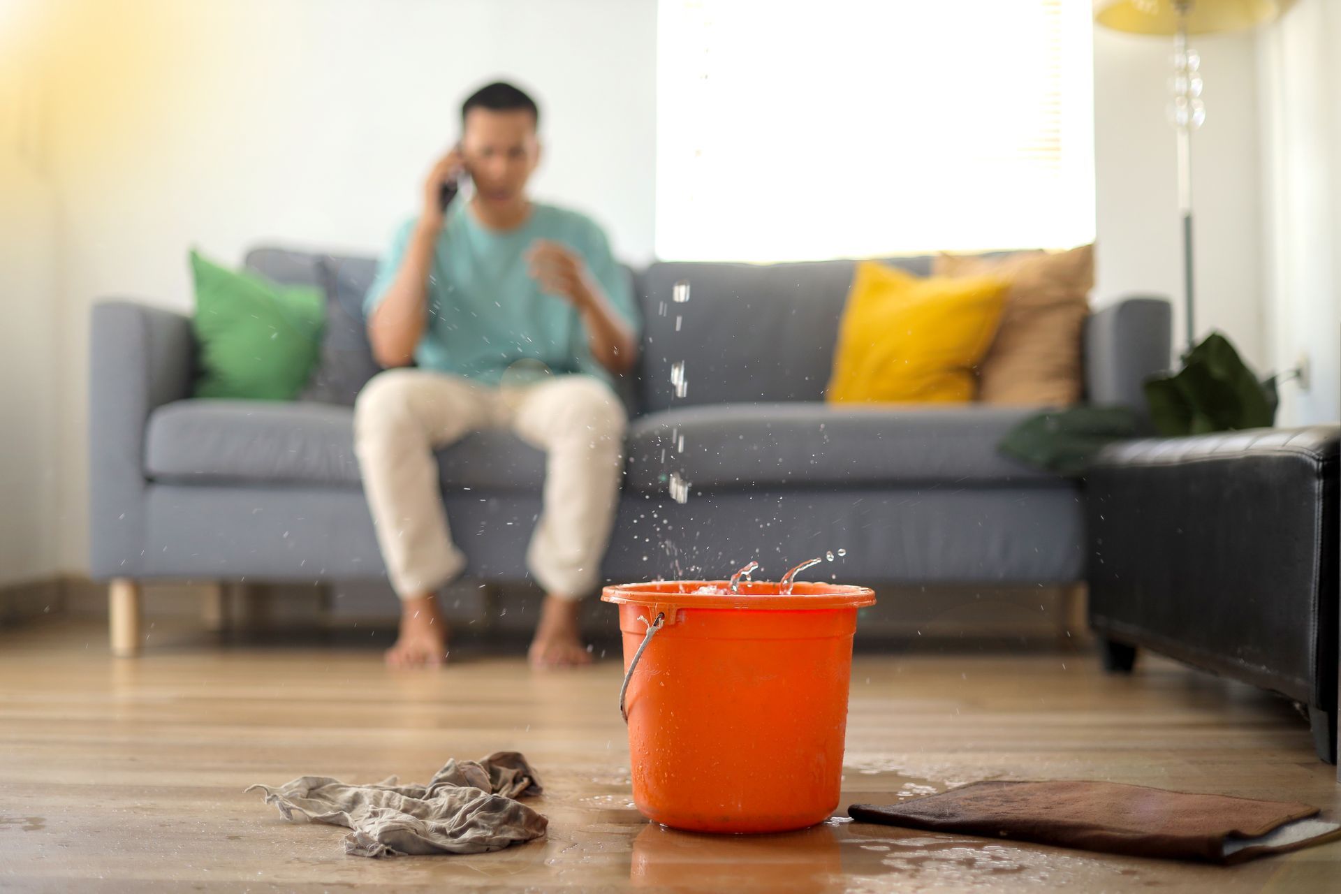 Orange bucket catching a leak on a wet floor, with a person sitting blurred on a couch in the background Orange bucket catching a leak on a wet floor, with a person sitting blurred on a couch in the background
