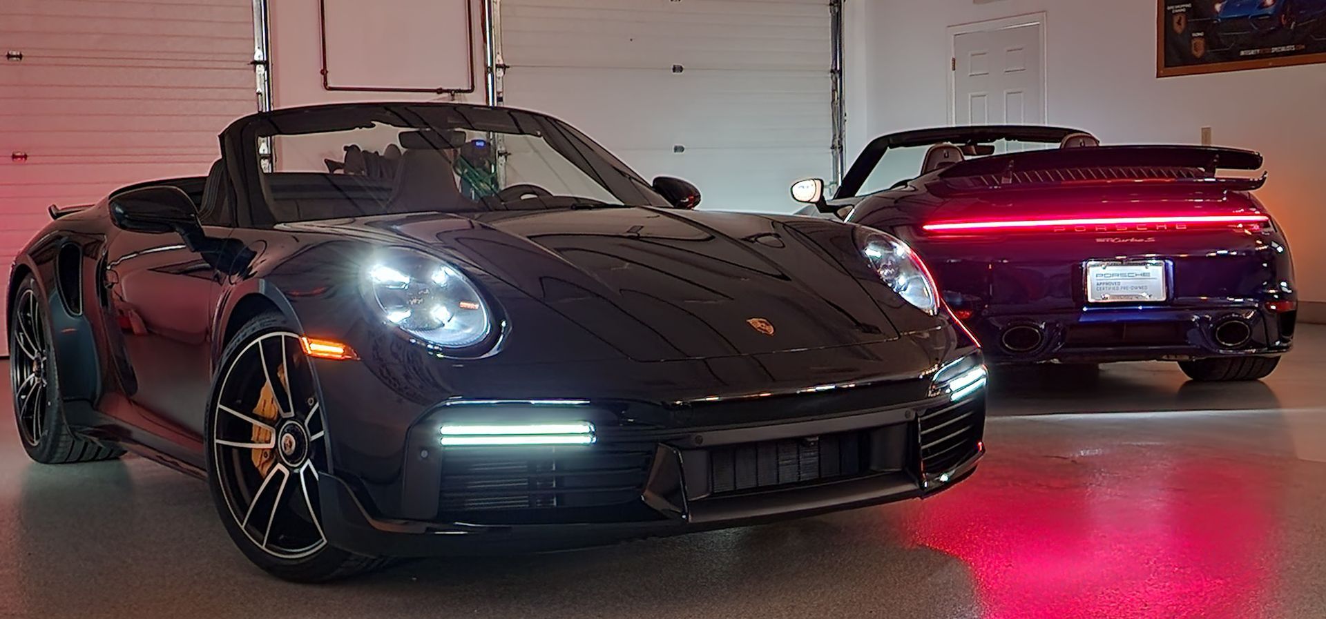 Two black Porsche 911 convertibles inside a garage, one in front and one in the back, under red lights.