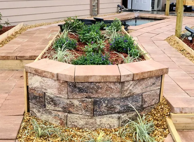 Curved brick planter box with colorful plants, framed by a red brick walkway, in an outdoor setting.