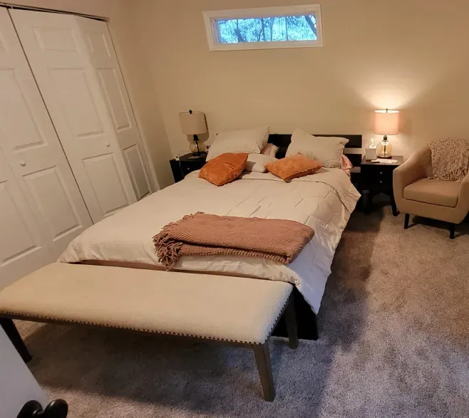 Bedroom with a bed, bench, closet, armchair, and lamps. Beige walls and bedding with orange accents.