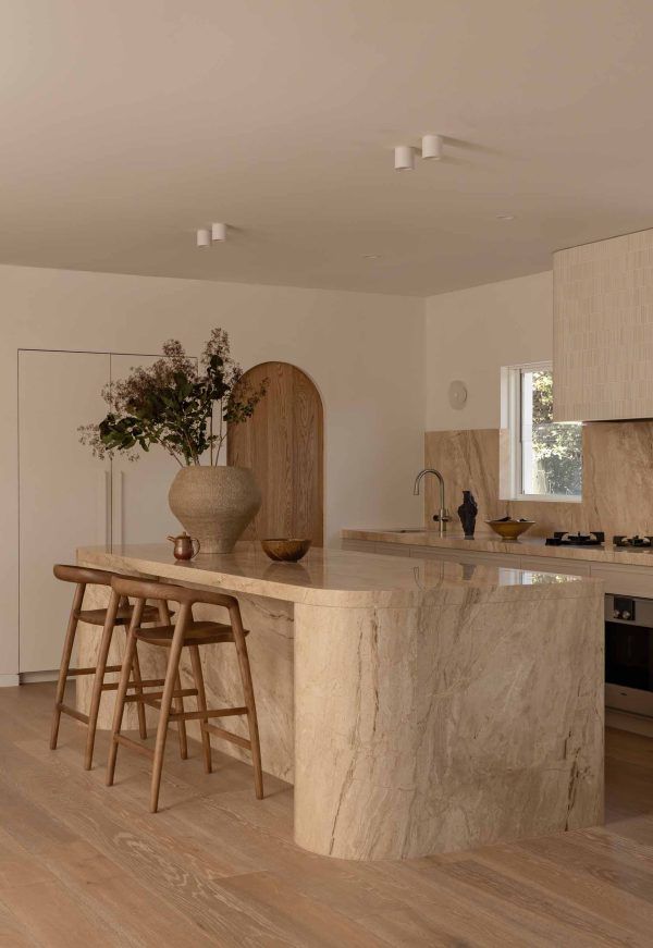 Warm minimalist kitchen with stone island, wooden stools, and neutral decor in a bright open room — Southeast Stone Pty Ltd In Caloundra West, QLD