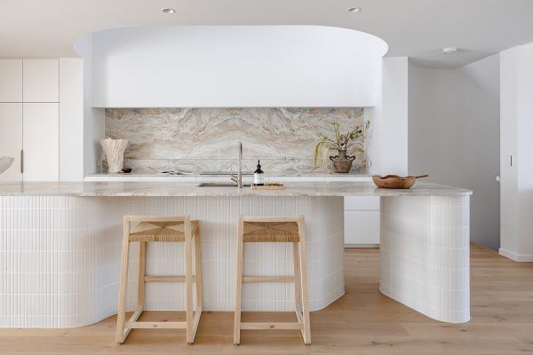 Minimalist white kitchen with curved island, two stools, and light wood floor — Southeast Stone Pty Ltd In Caloundra West, QLD