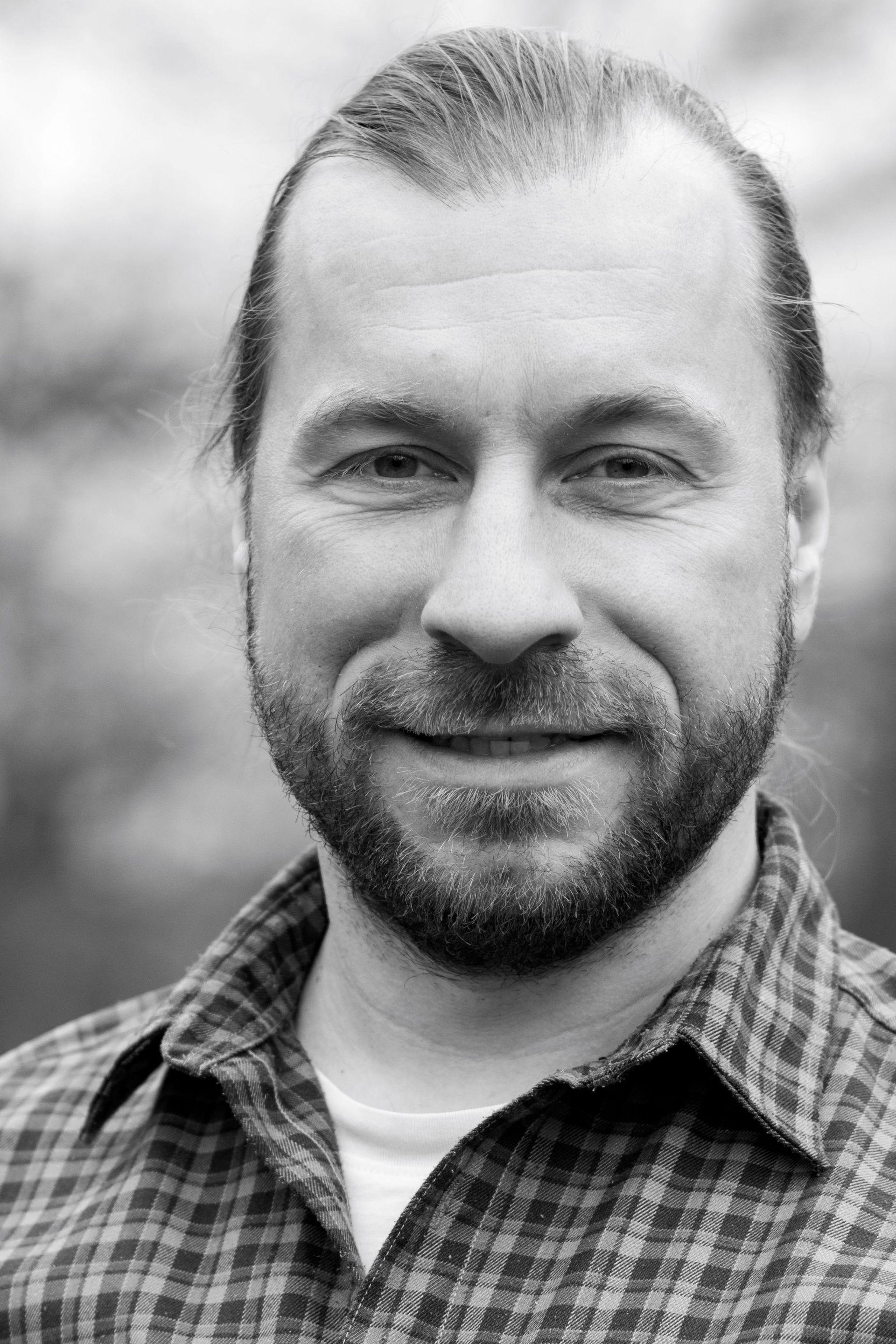 A man with a beard wearing a plaid shirt is smiling in a black and white photo.