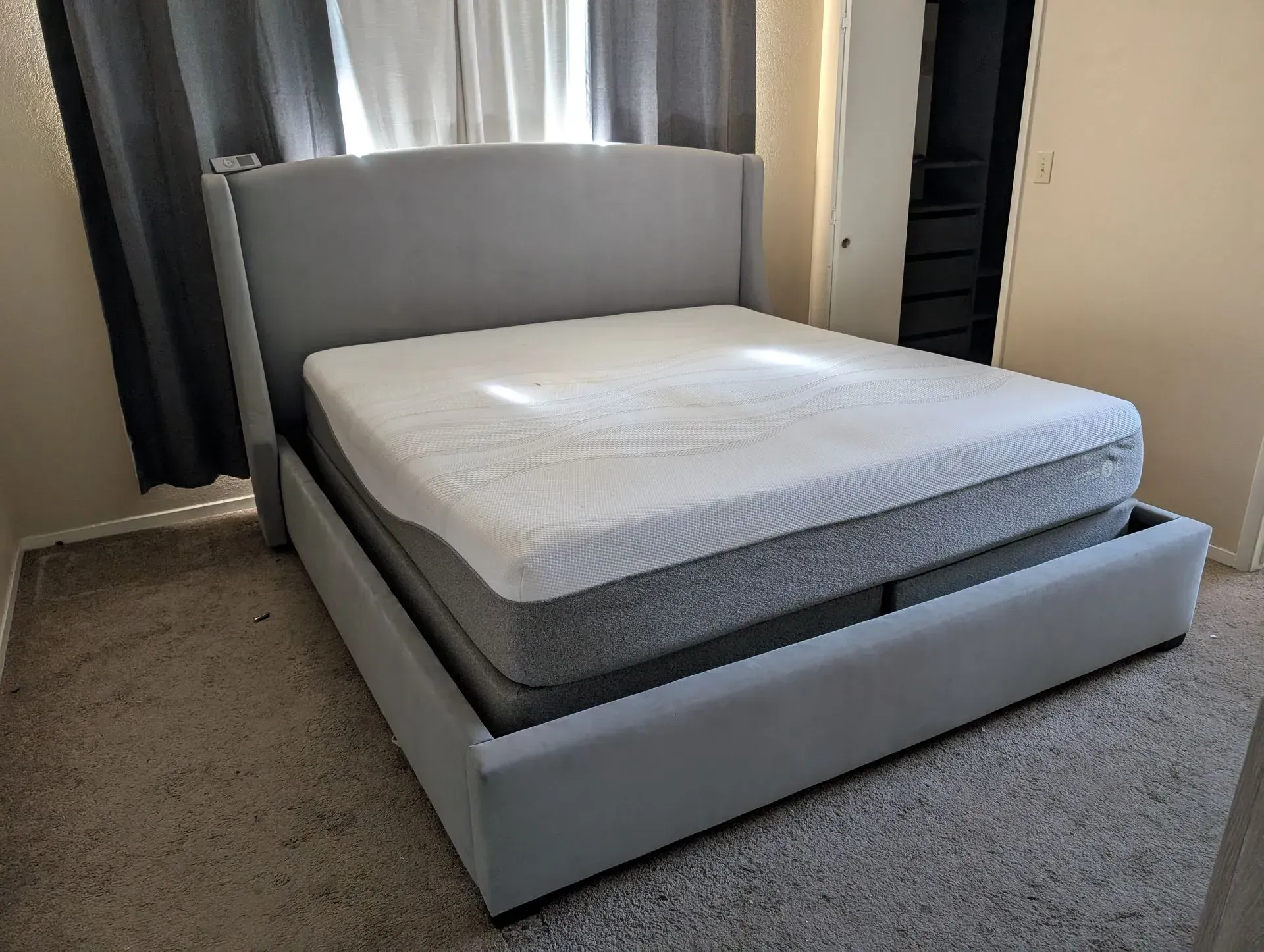 Modern gray upholstered bed with white mattress in a carpeted bedroom near a window.