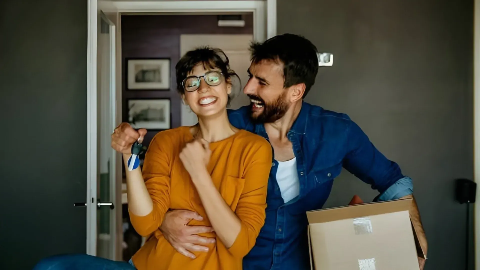 Two people smiling in a doorway, holding a moving box and keys in a home interior.