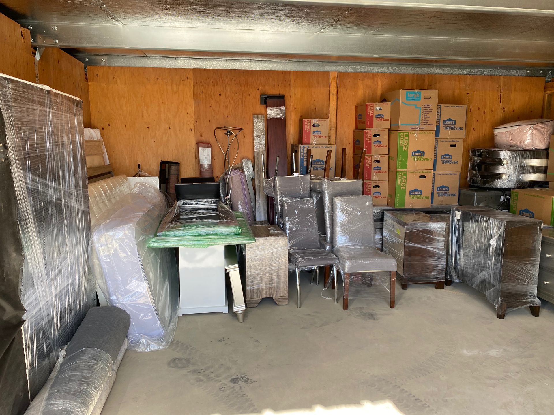 Storage room with stacked boxes, wrapped furniture, and metal chairs on a concrete floor.