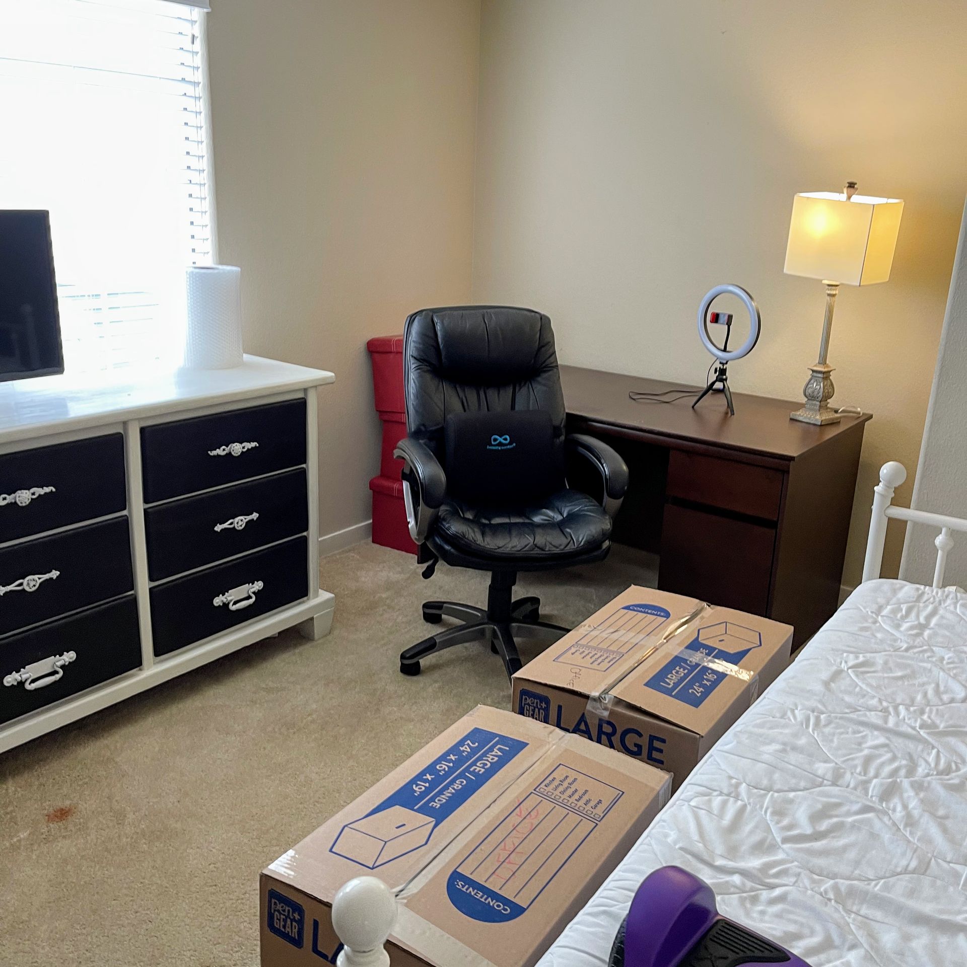 Neat bedroom with desk, lamp, dresser, and two cardboard boxes on the floor beside the bed.