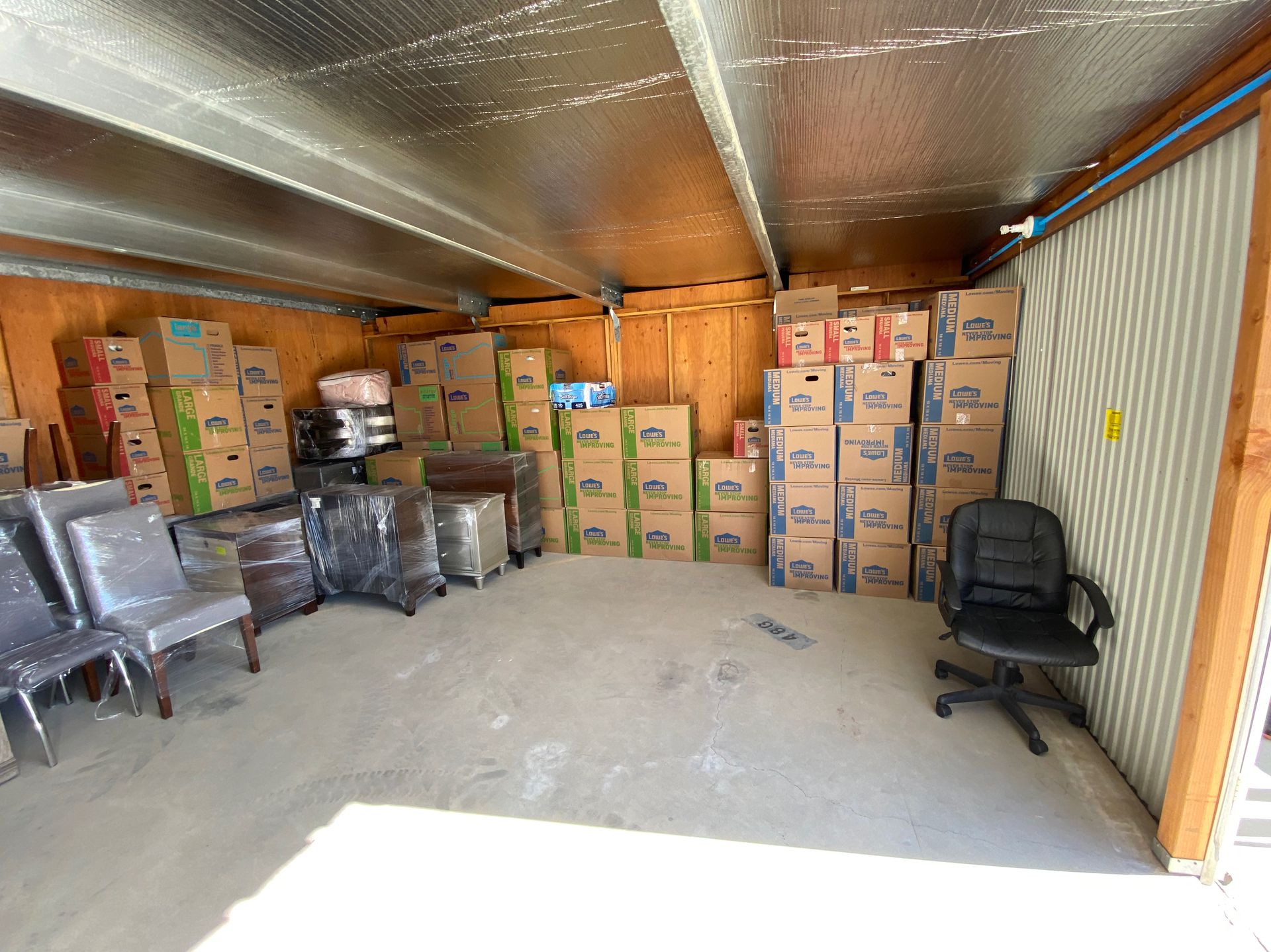 Storage unit interior with stacked cardboard boxes, plastic-wrapped items, and a black chair on the right