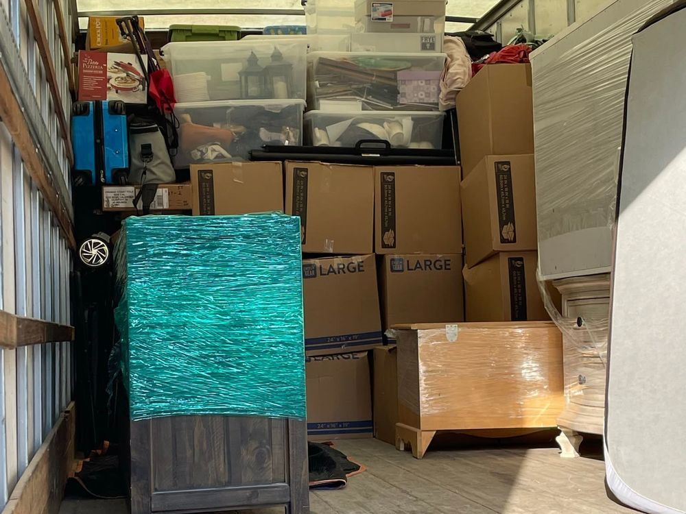 Storage area packed with stacked cardboard boxes, plastic bins, and wrapped furniture in a narrow garage space