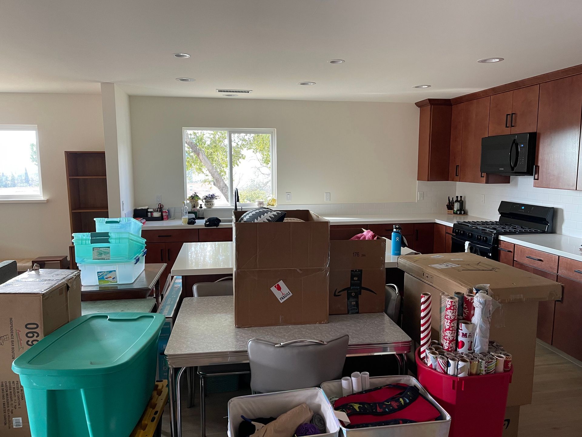 Packed living room with moving boxes, bins, and kitchen cabinets; sunlight through a window.