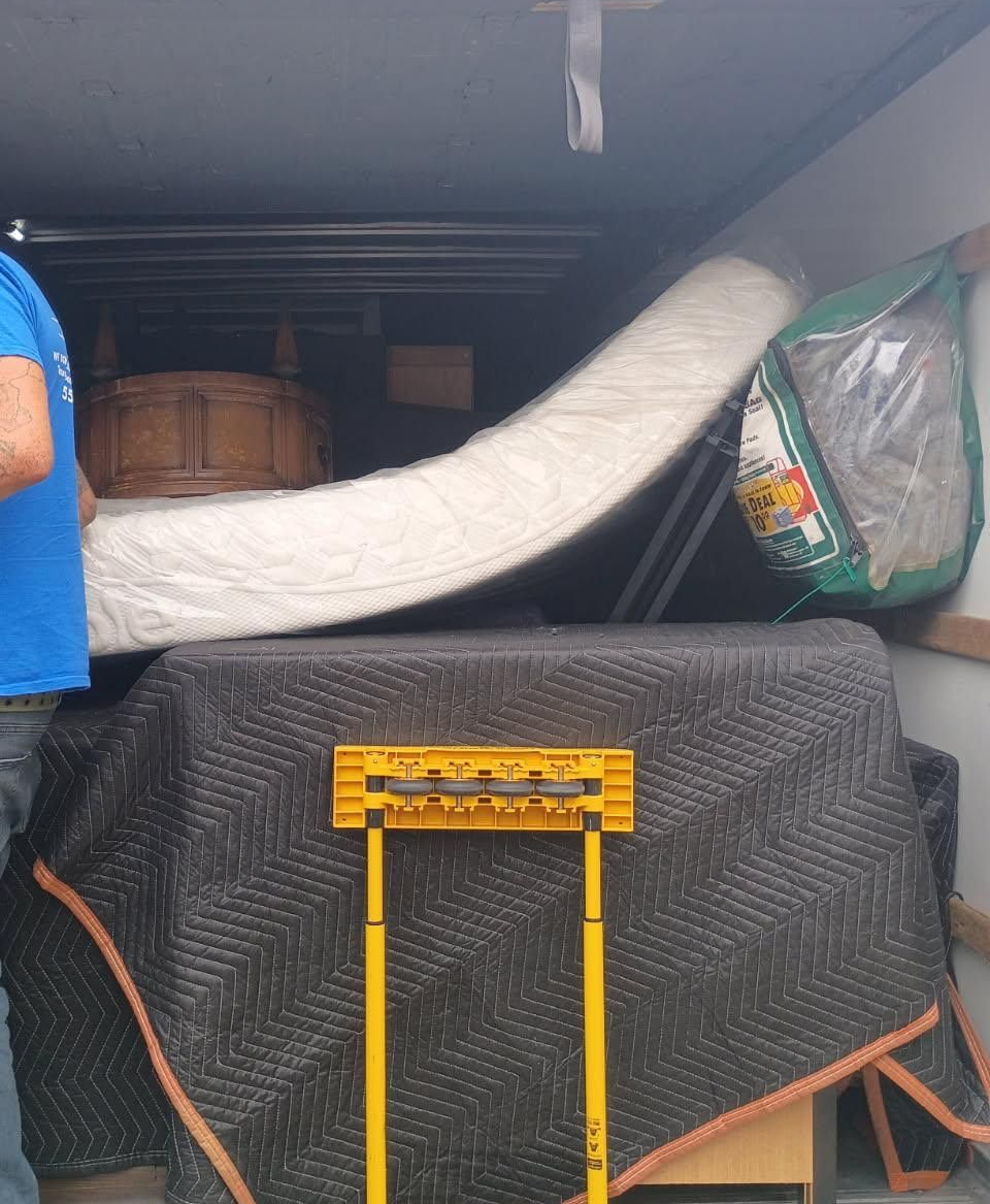 Packed storage area with a white rolled mattress, covered items, and a black netted cart in front.