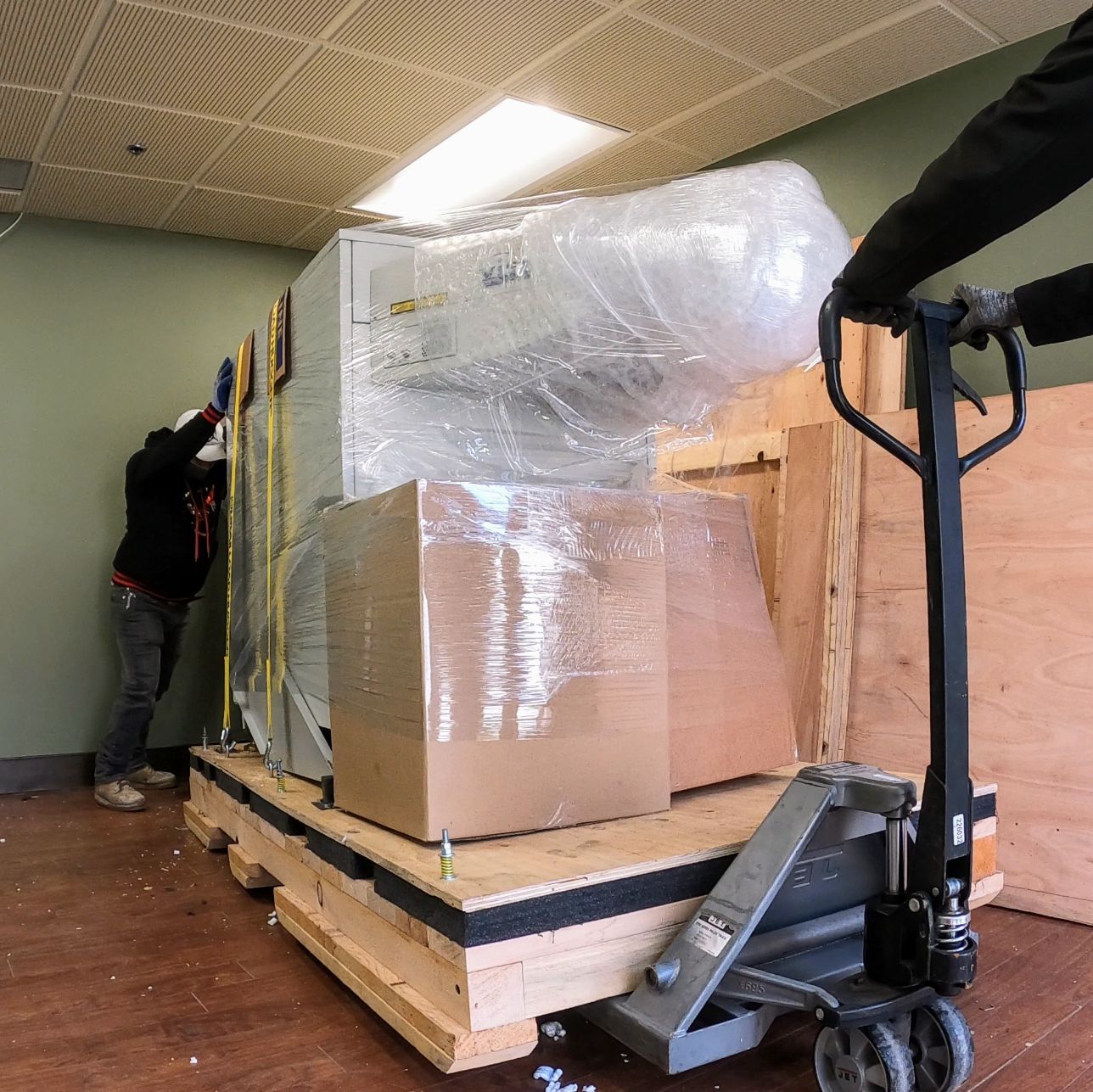 Packed furniture wrapped in plastic on a pallet, being moved with a hand truck in a room.
