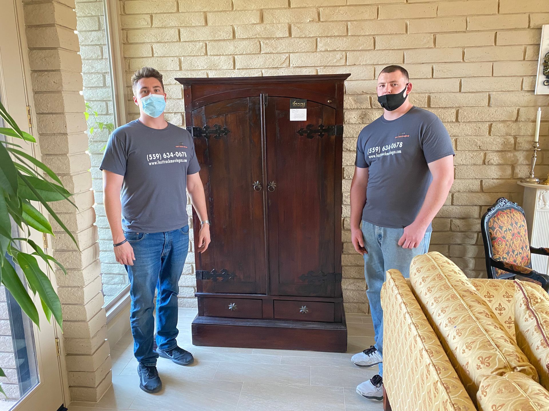 Two masked men standing beside a large wooden cabinet in a brick-walled living room