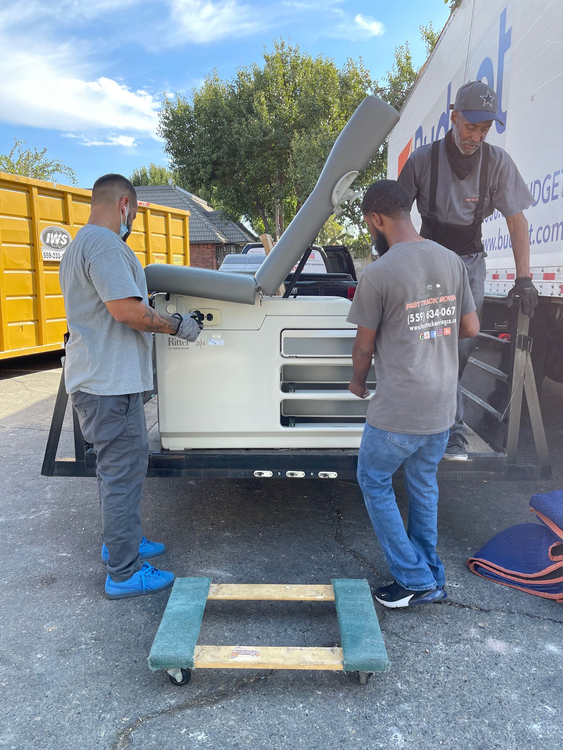 Workers loading a large white machine onto a trailer outdoors, with a dump truck in the background.