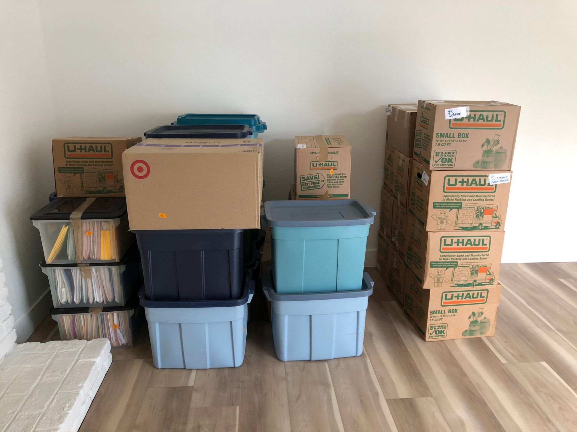 Stacked moving boxes and storage bins in a room with a wooden floor, including shelves of files on the left.