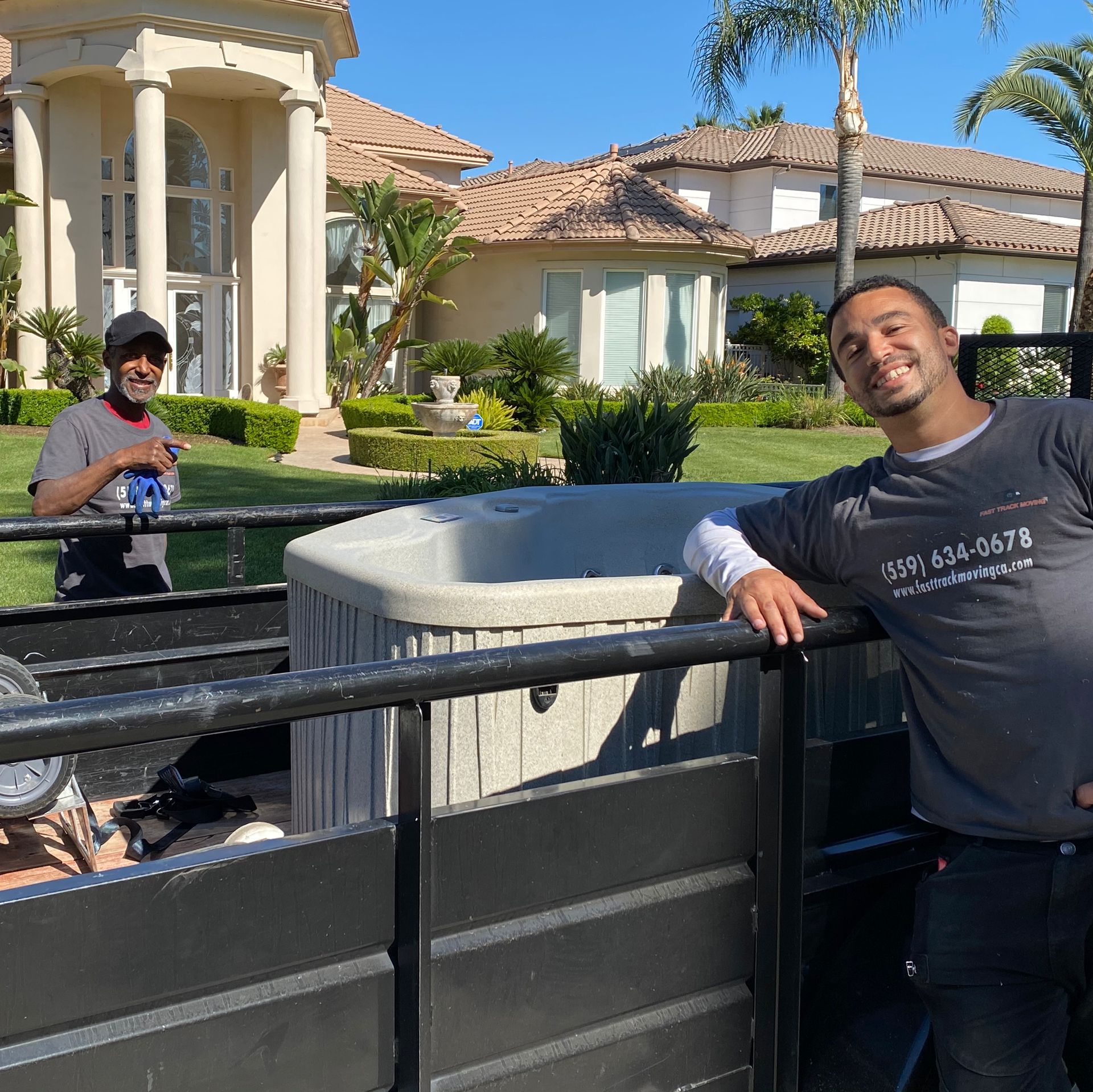 Two people posing by a black fence in front of a large house and palm trees on a sunny day