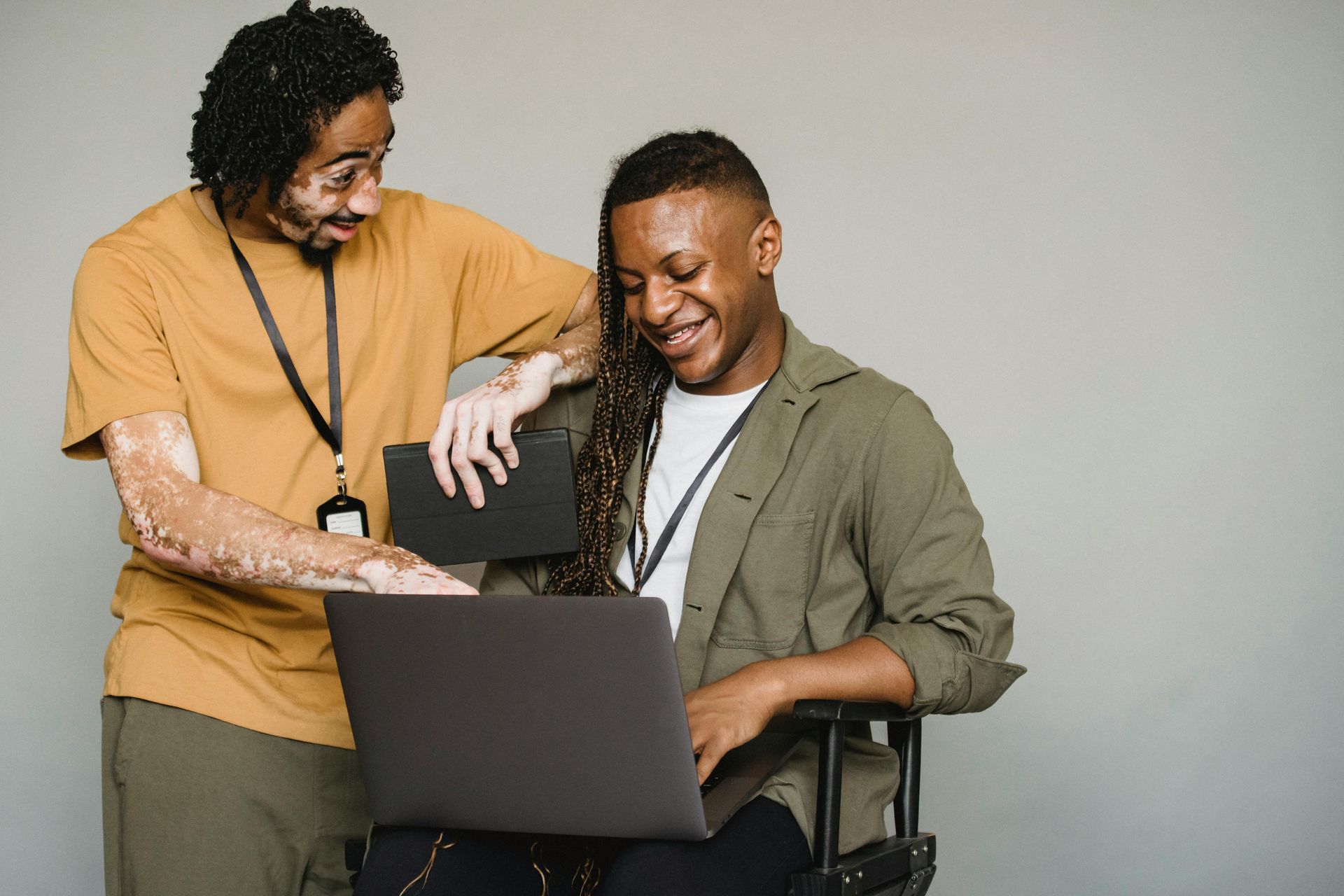 Dois homens olhando para um laptop, um deles com manchas de vitiligo. Sorrindo, fundo neutro.