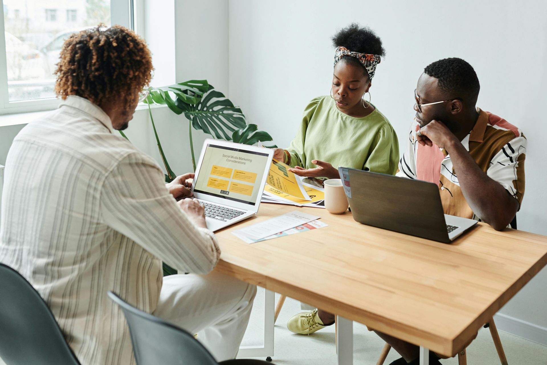 Três pessoas negras em uma mesa, discutindo trabalho. Uma usa um laptop, a outra olha papéis.