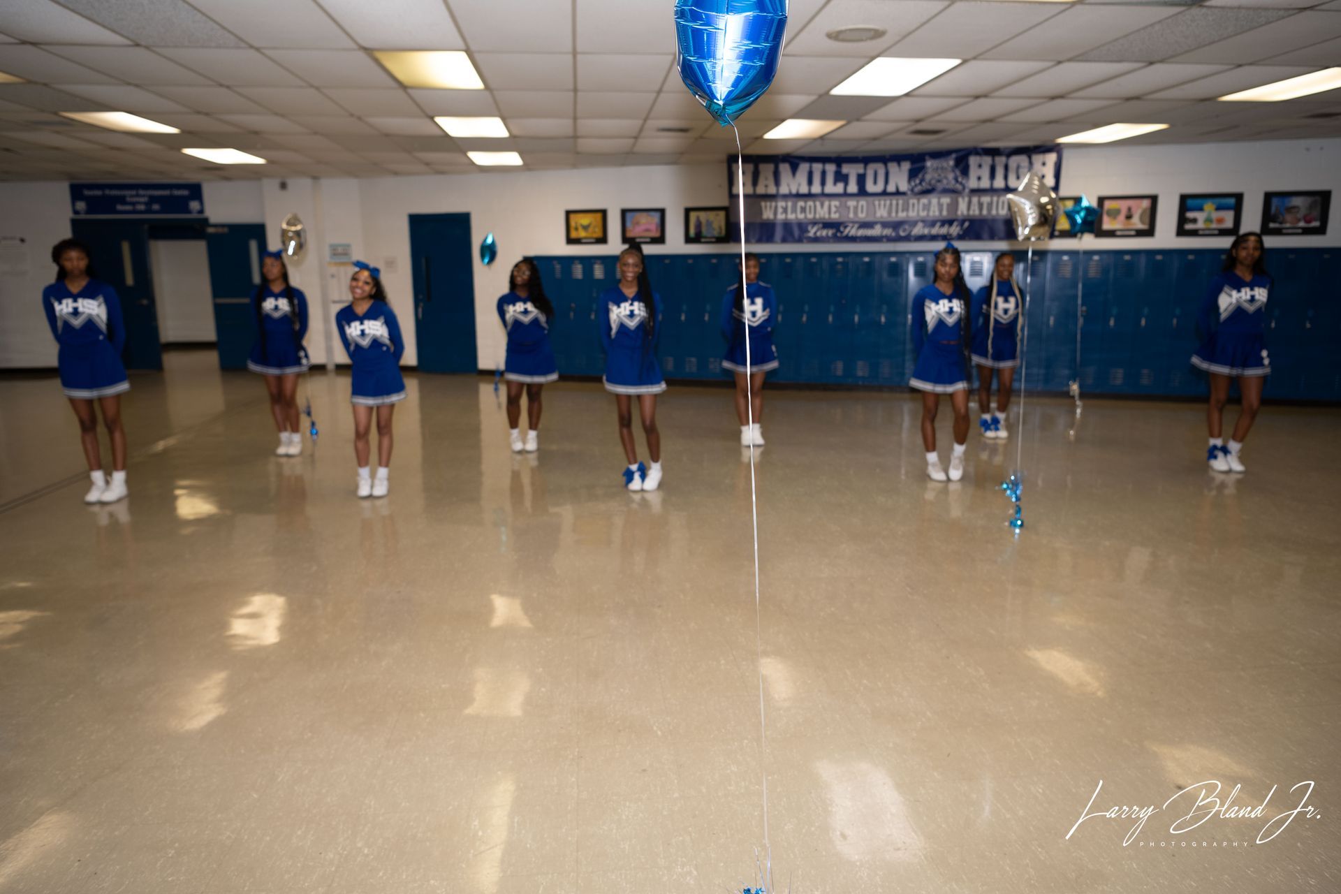 Cheerleaders in blue uniforms standing in a hallway under a balloon. 