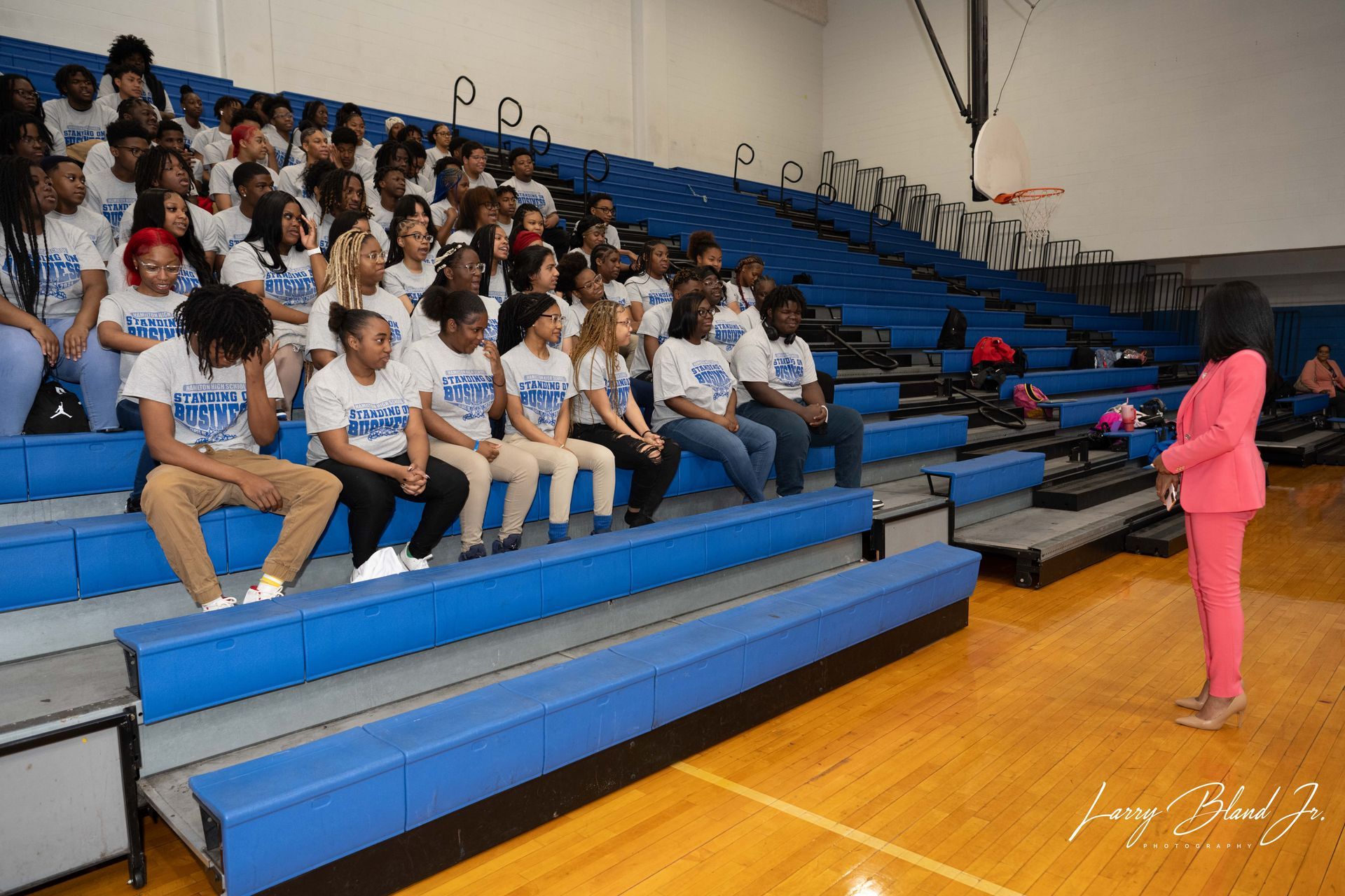 A woman in a pink suit speaks to a seated group of students in a gymnasium.