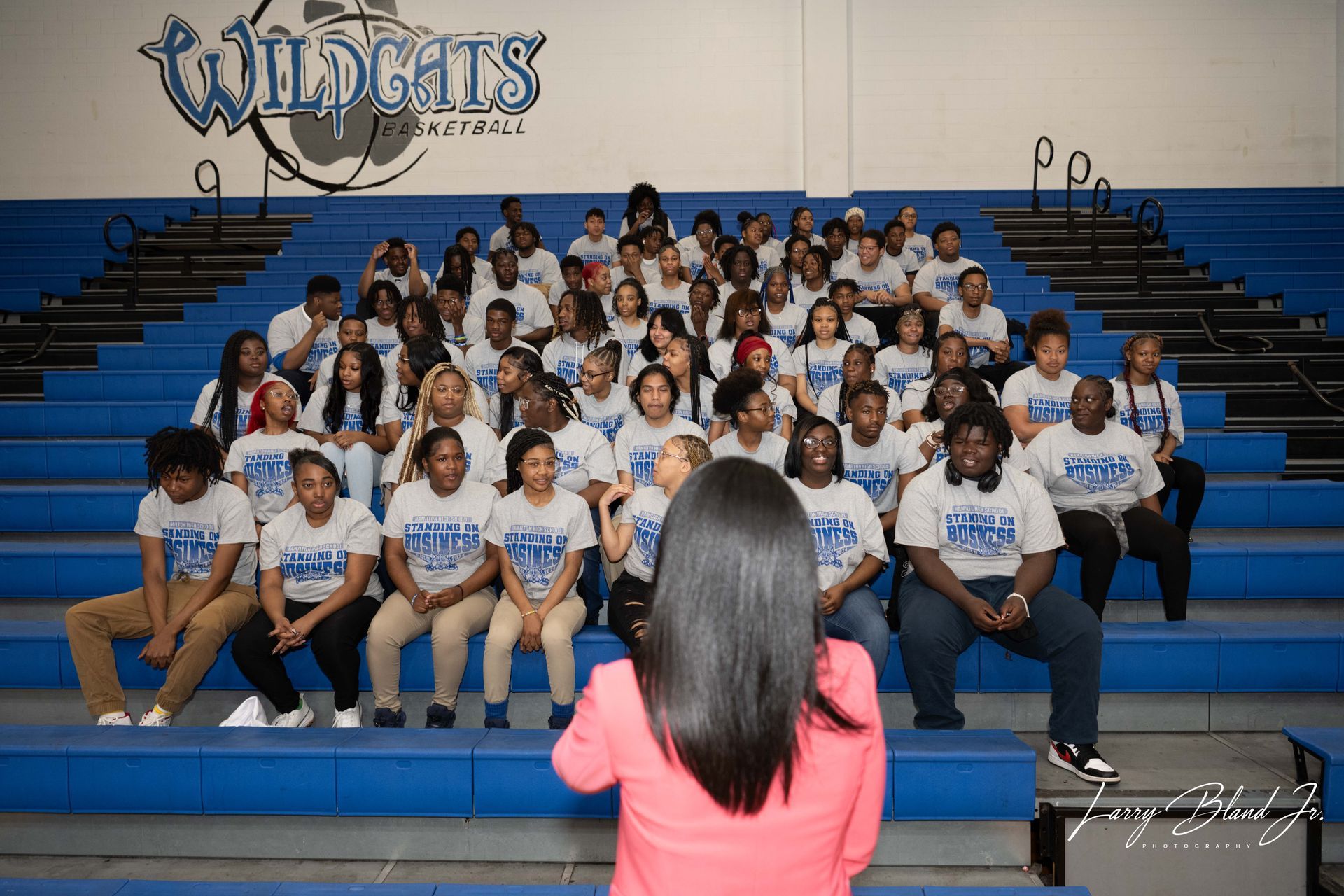 Group photo of students in blue and gray shirts on bleachers. Wildcats logo in background.