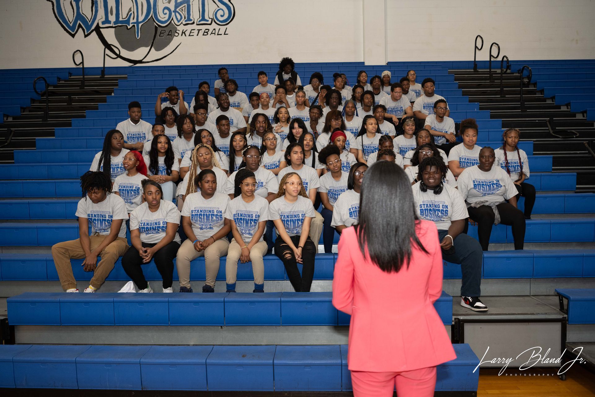 Group photo of people on bleachers in a basketball gym, wearing matching t-shirts, facing a person in a pink suit.