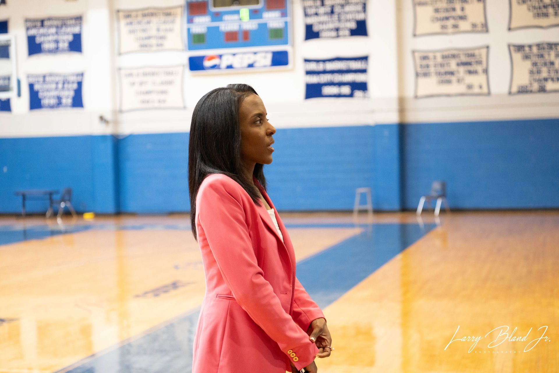 Woman in pink blazer standing in a gymnasium with a basketball court.