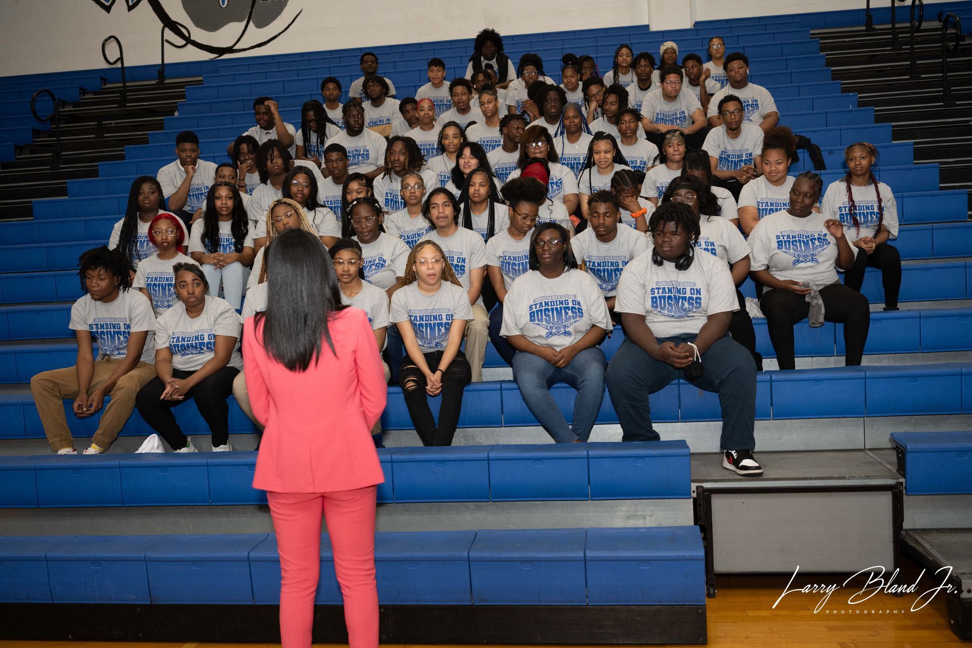 Woman in pink suit speaks to a group wearing matching shirts on blue bleachers.