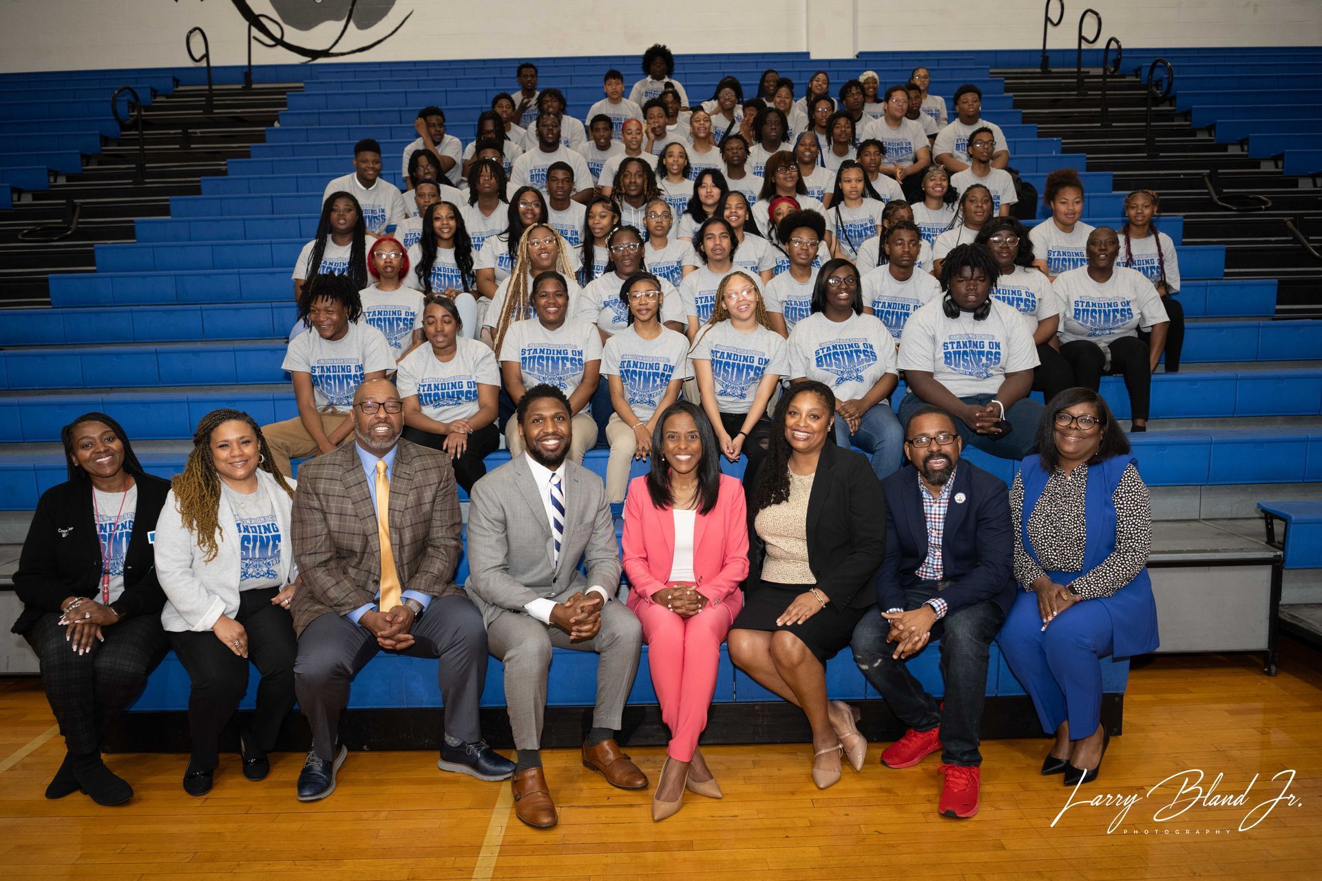 Group photo on blue bleachers; students wearing t-shirts, adults in business attire.